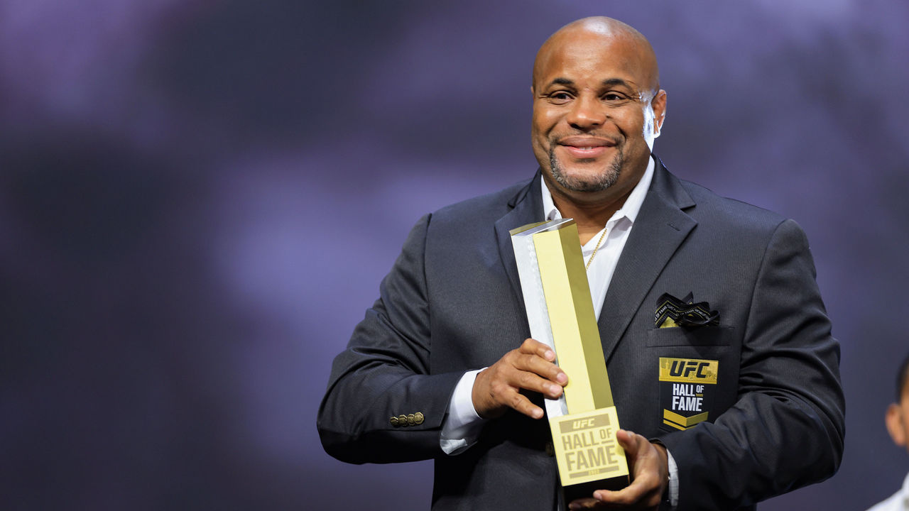 Daniel Cormier in a suit holds his UFC Hall of Fame trophy on stage
