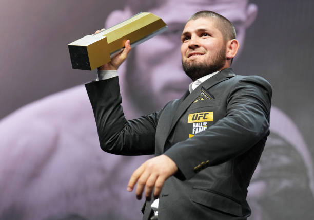 Khabib Nurmagomedov in a suit holds his UFC Hall of Fame trophy on stage